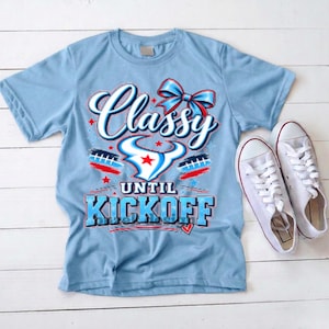 May include: A light blue short-sleeve t-shirt featuring a graphic print with a red, white, and blue bow, a stylized bull horn logo with a red star, and the text Classy UNTIL KICKOFF. A pair of white canvas sneakers are placed next to the shirt.