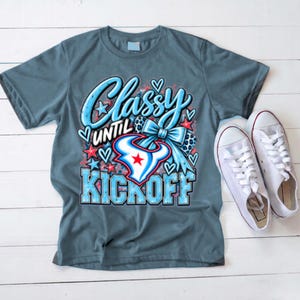 May include: A slate blue short-sleeve t-shirt featuring a graphic print with the text Classy UNTIL KICKOFF in light blue and white. The design includes a blue bow, red and blue stars, hearts, and a bull horn logo with a red star in the center.