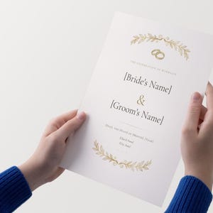 May include: A white wedding invitation card featuring gold foil illustrations of two interlocking rings and laurel leaf branches at the top and bottom. The text includes The Celebration of Marriage, Bride's Name, Groom's Name, Date, and location details.