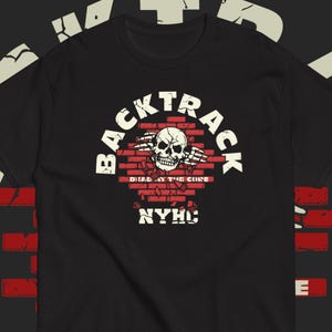 Op de afbeelding: Een zwart t-shirt met korte mouwen en een afbeelding van een witte schedel die door een rode bakstenen muur breekt. De tekst BACKTRACK staat in een boog erboven, met DEAD AT THE CORE in het midden en NYHC onderaan in een verweerd wit lettertype.