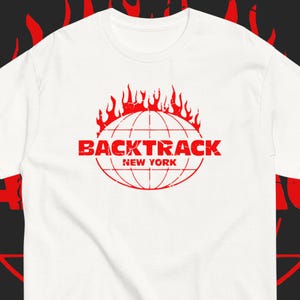 May include: A white short-sleeve t-shirt featuring a red graphic print of a globe with flames rising from the top. The text BACKTRACK NEW YORK is printed in a distressed red font across the center of the globe design on the front of the shirt.