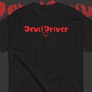 May include: A black short-sleeved t-shirt featuring the text DevilDriver printed in a distressed red font across the chest. Below the text is a small red graphic symbol. The design is centered on the front of the garment with a crew neckline.