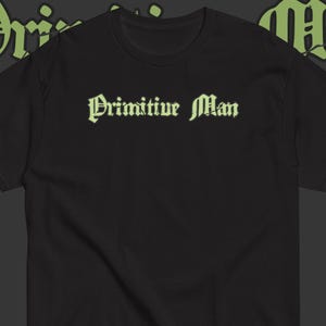May include: A black short-sleeved t-shirt featuring the text "Primitive Man" printed in a light green, gothic-style font across the chest. The shirt has a crew neckline and a simple, solid black design with the text centered on the front.