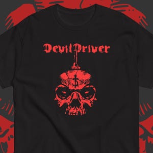 Może przedstawiać: Czarny t-shirt z krótkim rękawem z czerwonym nadrukiem graficznym na klatce piersiowej. Wzór przedstawia napis DevilDriver stylizowaną czcionką nad szczegółową ilustracją czaszki z symbolem krzyża na czole i kolcem na górze.
