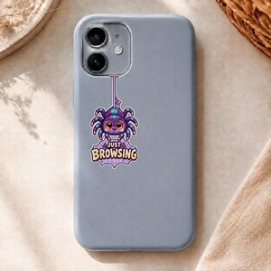 Könnte beinhalten: Eine hellgraue Smartphone-Hülle mit einem Aufkleber einer niedlichen lila-blauen Spinne, die an einem Netz hängt. Die Spinne hat große braune Augen und ein kleines Lächeln. Unter der Spinne steht der Text JUST BROWSING in fetten gelben Buchstaben.