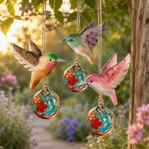 Könnte beinhalten: Drei handgeschnitzte Holz-Kolibri-Ornamente in Rosa-, Grün- und Brauntönen hängen an Schnüren. Jeder Vogel ist über einer Glaskugel mit roten Blumendesigns und wirbelnden blau-grünen Mustern aufgehängt. Die Ornamente werden im Freien ausgestellt.