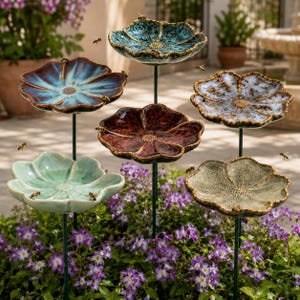 以下が含まれることがあります： 細い金属製の杭に取り付けられた6つの花形の陶器製バードバスのコレクション。それぞれがティール、ブルー、ダークレッド、ホワイト、ベージュの色合いのユニークな釉薬仕上げを特徴とし、紫色の花の間でさまざまな高さに配置されています。