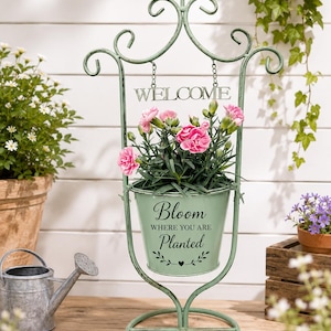 Op de afbeelding: Een decoratieve, mintgroene metalen plantenstandaard met een vogel erop, met een hangend bord met de tekst "WELCOME". De standaard bevat een groene emmer met roze bloemen en de tekst "Bloom where you are planted".