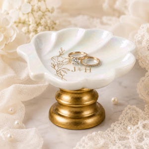 May include: A white iridescent shell-shaped ring dish with a gold pedestal base. The dish features a gold floral illustration and the text J+H. Two gold rings, one with a central stone and one with a pavé band, rest inside the dish.