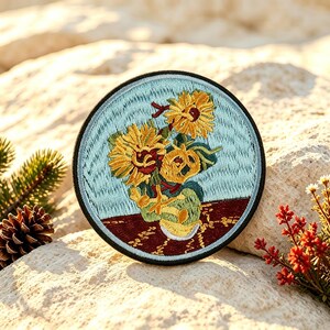 Könnte beinhalten: Ein kreisförmiger bestickter Aufnäher mit einer lebendigen Darstellung gelber Sonnenblumen in einer Vase vor hellblauem Hintergrund auf einer dunkelroten Fläche. Das Motiv ist von einem dicken schwarzen Rand umgeben.