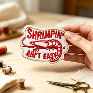 Puede incluir: Un parche bordado con fondo blanco y borde dorado. Presenta una ilustración de un camarón rojo en el centro con el texto rojo SHRIMPIN' arriba y AIN'T EASY debajo del camarón, todo en una fuente llamativa y estilizada.