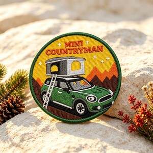 Peut inclure: Un écusson brodé circulaire représentant une voiture Mini Countryman verte avec une tente de toit et une échelle. Le fond montre des montagnes orange et un ciel jaune avec des étoiles blanches. Le texte MINI COUNTRYMAN est brodé en rouge.