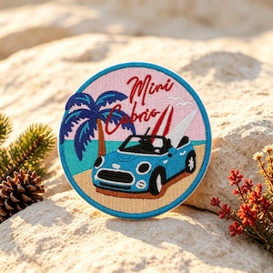 Pode incluir: Um emblema bordado circular apresentando um carro conversível azul claro em uma praia de areia com uma palmeira azul escura e pranchas de surf vermelhas e brancas. O texto Mini Cabrio está bordado em vermelho acima do carro num céu rosa.