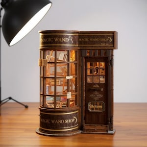 Peut inclure: Une maquette miniature détaillée d'une boutique de baguettes magiques. La boutique présente une vitrine ronde remplie de livres et une porte en bois avec le mot "Welcome". L'enseigne de la boutique indique "Magic Wand Shop".
