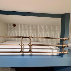May include: A horizontal safety railing constructed from metallic bronze-toned industrial pipes and fittings. The structure features three parallel bars connected by vertical segments, installed as a protective barrier for a lofted sleeping area.