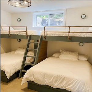 May include: A custom-built bunk bed structure featuring two lower beds and two upper beds with dark green frames. The upper bunks include copper-toned metal pipe railings. A central wooden ladder connects the levels, and white bedding is used.