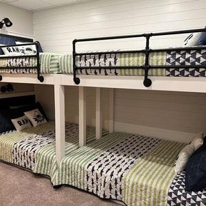 May include: A white wooden bunk bed frame with black industrial pipe railings. The beds feature quilted bedding with green and white stripes, a shark pattern, and navy blue geometric accents. White throw pillows with the text RAH are included.