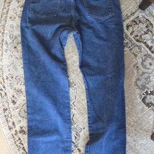 May include: A pair of blue denim jeans featuring a classic five-pocket design, a waistband with belt loops, and a tan leather patch on the back right pocket. The back pockets are stitched with a signature W pattern in a darker blue thread.