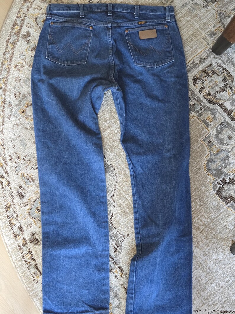 May include: A pair of blue denim jeans featuring a classic five-pocket design, a waistband with belt loops, and a tan leather patch on the back right pocket. The back pockets are stitched with a signature W pattern in a darker blue thread.