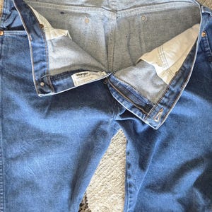 May include: A pair of blue denim jeans featuring a metal zipper fly, copper-toned rivets, and a white interior waistband label with the Wrangler brand name. The jeans are shown from the front with the fly open, revealing white pocket lining.