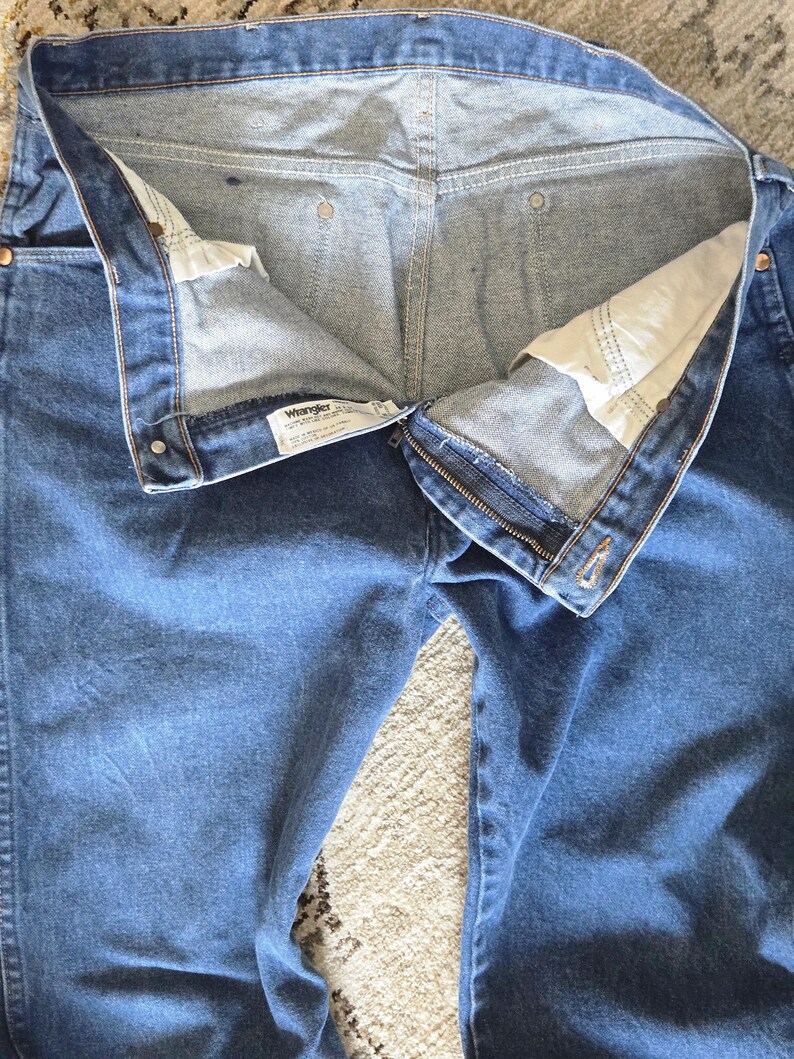 May include: A pair of blue denim jeans featuring a metal zipper fly, copper-toned rivets, and a white interior waistband label with the Wrangler brand name. The jeans are shown from the front with the fly open, revealing white pocket lining.