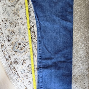 May include: A pair of dark blue denim jeans featuring a straight leg cut and visible stitching along the side seam. A yellow measuring tape is placed alongside the leg, indicating a length of approximately 31 inches from the top to the hem.