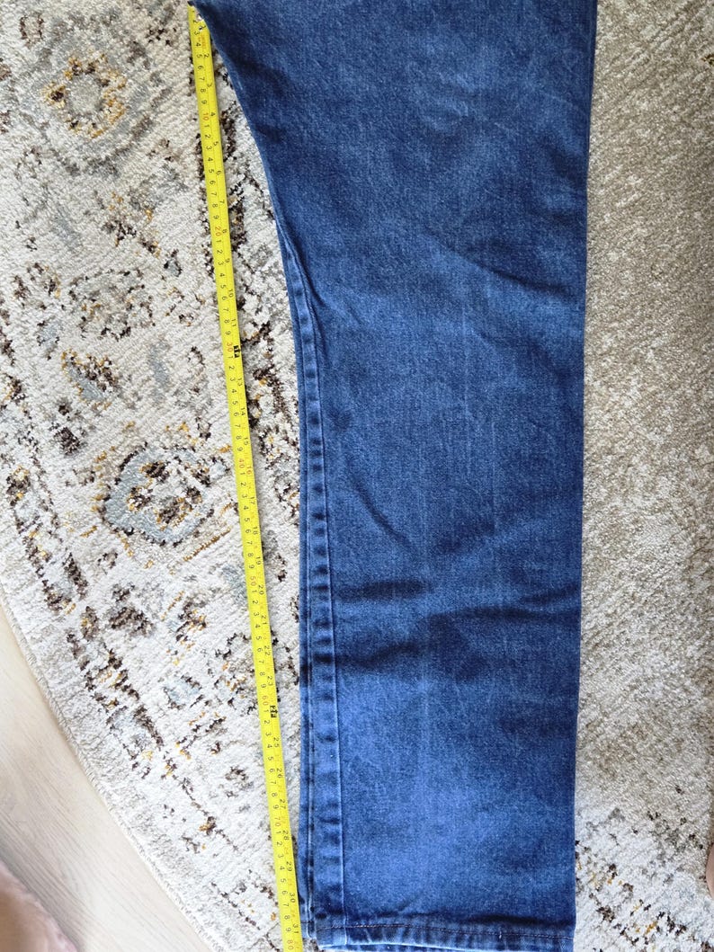 May include: A pair of dark blue denim jeans featuring a straight leg cut and visible stitching along the side seam. A yellow measuring tape is placed alongside the leg, indicating a length of approximately 31 inches from the top to the hem.