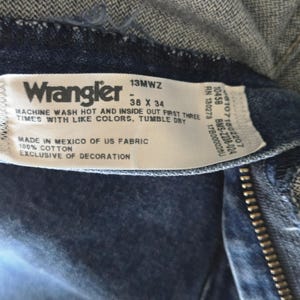 May include: A white fabric clothing tag sewn into dark blue denim jeans. The tag features the Wrangler logo, the style number 13MWZ, and the size 38 X 34. It includes care instructions, states 100% cotton, and indicates it was made in Mexico of US fabric.