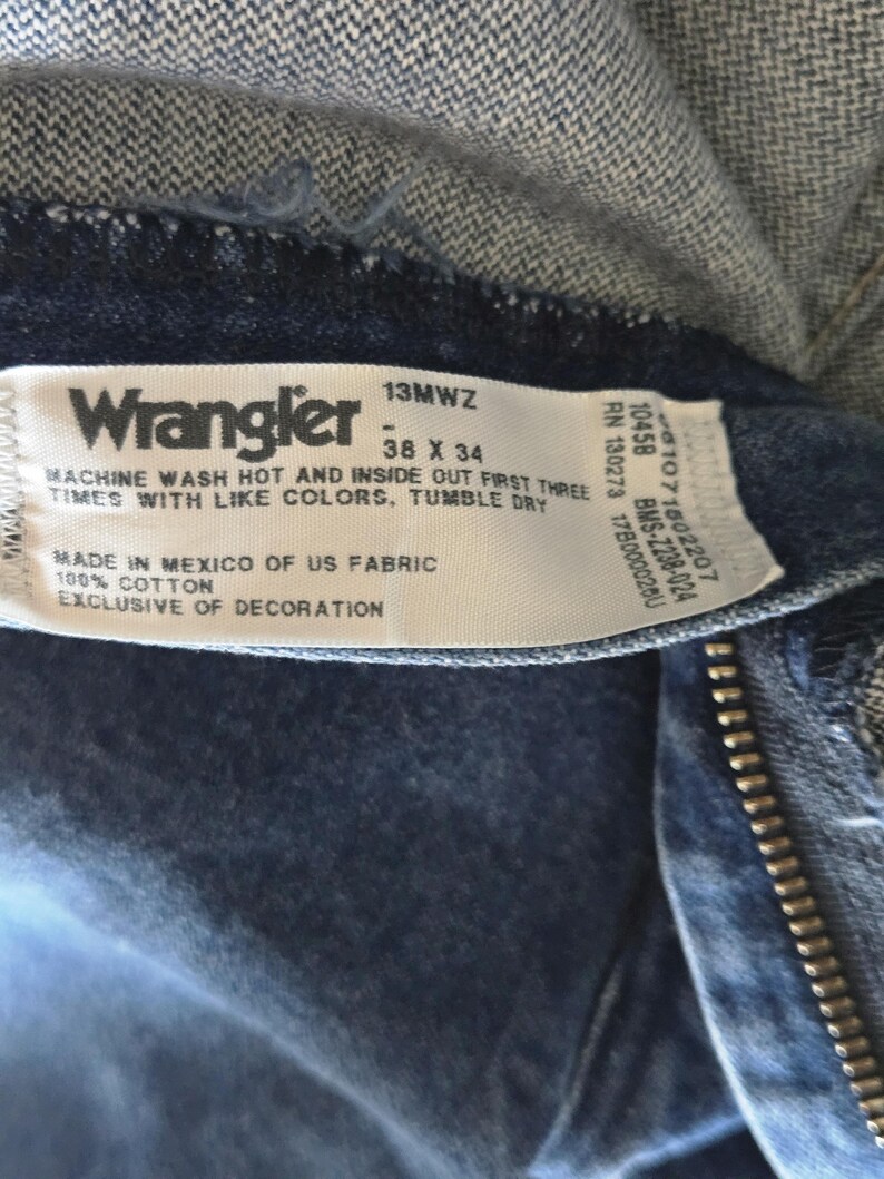 May include: A white fabric clothing tag sewn into dark blue denim jeans. The tag features the Wrangler logo, the style number 13MWZ, and the size 38 X 34. It includes care instructions, states 100% cotton, and indicates it was made in Mexico of US fabric.