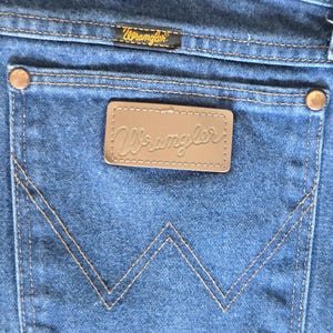 May include: Blue denim jeans featuring a back pocket with a signature W-shaped stitch pattern, copper-colored metal rivets at the corners, and a rectangular brown leather patch embossed with the Wrangler logo above the pocket.