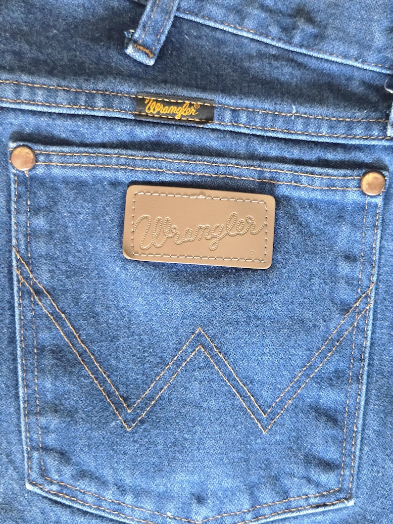 May include: Blue denim jeans featuring a back pocket with a signature W-shaped stitch pattern, copper-colored metal rivets at the corners, and a rectangular brown leather patch embossed with the Wrangler logo above the pocket.