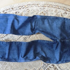 May include: A pair of medium-wash blue denim jeans featuring a classic five-pocket design, a button fly closure, and visible copper-toned rivets. The jeans have a straight-leg cut with traditional topstitching along the seams and waistband.