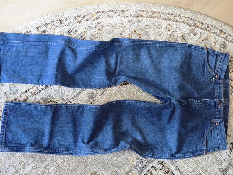 May include: A pair of medium-wash blue denim jeans featuring a classic five-pocket design, a button fly closure, and visible copper-toned rivets. The jeans have a straight-leg cut with traditional topstitching along the seams and waistband.