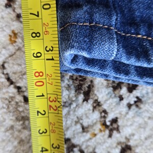 May include: A dark blue denim fabric with visible orange stitching along the hem. A yellow measuring tape is positioned vertically next to the fabric, showing a measurement extending from approximately 29.5 inches to 33.5 inches in length.