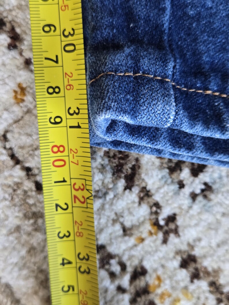 May include: A dark blue denim fabric with visible orange stitching along the hem. A yellow measuring tape is positioned vertically next to the fabric, showing a measurement extending from approximately 29.5 inches to 33.5 inches in length.