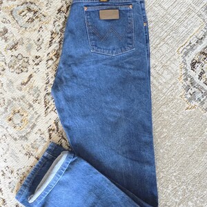 May include: Medium blue denim jeans featuring a classic five-pocket design, a button and zipper fly closure, and a brown leather patch on the back pocket with the Wrangler brand logo and a stitched W pattern on the rear pocket fabric.