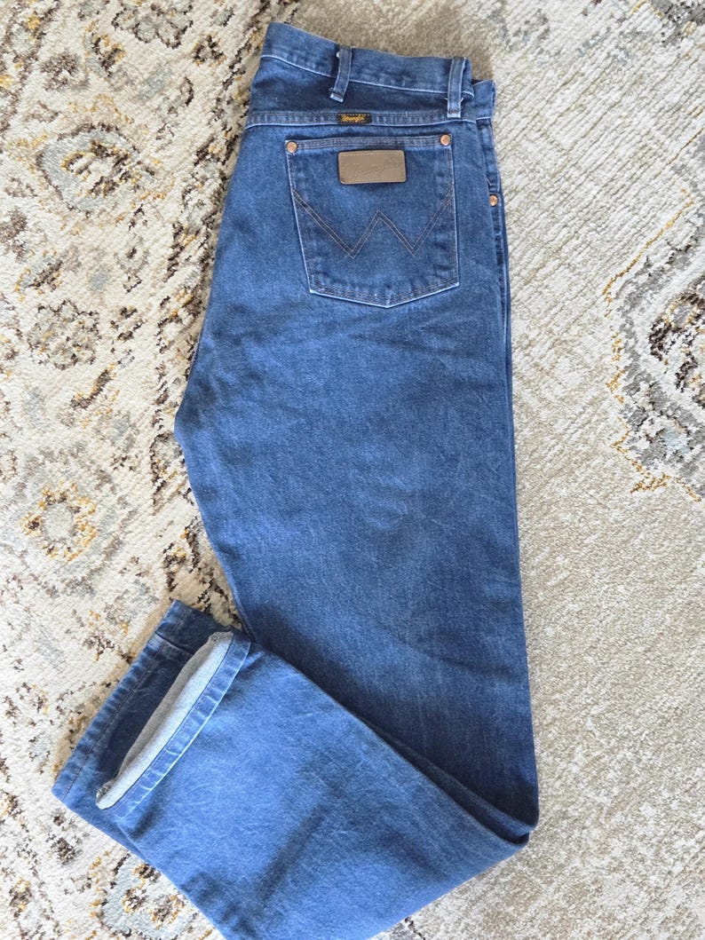 May include: Medium blue denim jeans featuring a classic five-pocket design, a button and zipper fly closure, and a brown leather patch on the back pocket with the Wrangler brand logo and a stitched W pattern on the rear pocket fabric.