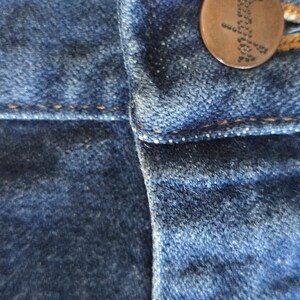 May include: A close-up view of blue denim fabric featuring a dark brown metal button at the waistband. The button is embossed with the word Wrangler in a dotted font. The denim has visible orange stitching along the seams and a textured surface.