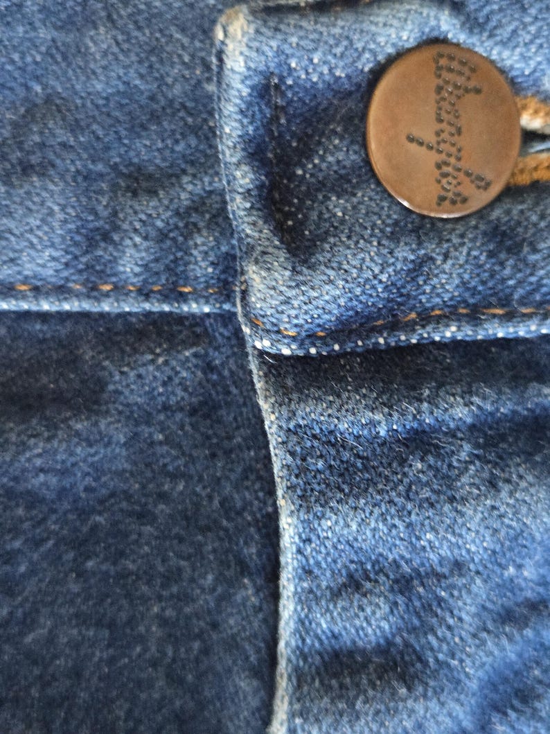 May include: A close-up view of blue denim fabric featuring a dark brown metal button at the waistband. The button is embossed with the word Wrangler in a dotted font. The denim has visible orange stitching along the seams and a textured surface.