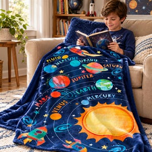 May include: A dark blue plush blanket featuring a colorful solar system print with the sun, planets labeled Mercury, Venus, Earth, Mars, Jupiter, Saturn, Uranus, Neptune, and Pluto, along with rocket ship illustrations and yellow stars.