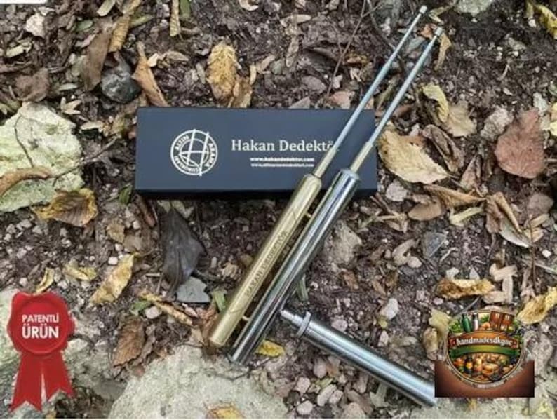 May include: Two metal detecting rods with gold and silver finishes, along with a black box with the text "Hakan Dedekt&ouml;r" and a logo. The image also includes a red seal with Turkish text and a decorative logo.