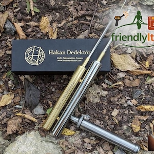 Puede incluir: Un juego de detectores de metales con dos varillas doradas y plateadas, una caja negra con letras doradas y una cinta roja con texto turco. La imagen está sobre un lecho de hojas y tierra.