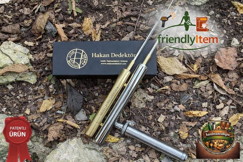 May include: A metal detector set with two gold and silver rods, a black box with gold lettering, and a red ribbon with Turkish text. The image is set on a bed of leaves and dirt.