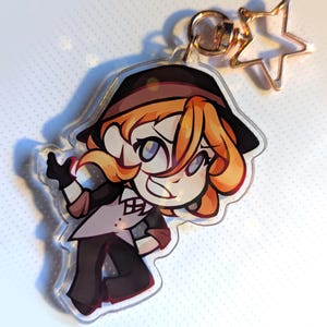 Peut inclure: Un porte-clés en acrylique transparent représentant un personnage de dessin animé aux cheveux roux, un chapeau noir et une tenue noire. Le porte-clés est doté d'un fermoir en forme d'étoile de couleur or et d'un porte-clés de couleur or.