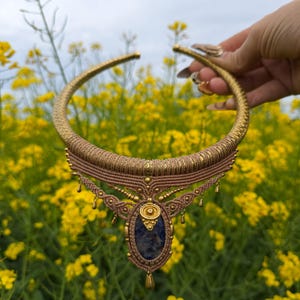 May include: A handcrafted gold-toned choker necklace with a central pendant featuring a blue stone. The necklace has intricate macrame detailing in shades of brown and gold, with small dangling accents. The background is a field of yellow flowers.