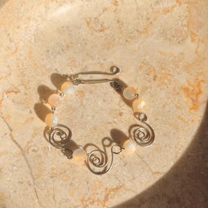 Op de afbeelding: Een armband met perzik kleurige kralen en zilverkleurige metalen accenten. De armband heeft een sluiting en decoratieve krullen. De sieraden worden getoond op een lichtgekleurd, getextureerd oppervlak.