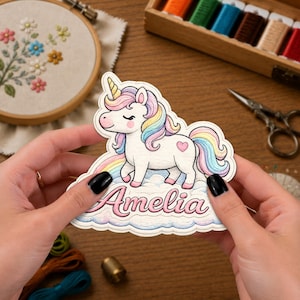 Puede incluir: Una pegatina troquelada que muestra un unicornio blanco con crin y cola de arcoíris pastel, un cuerno amarillo y un corazón rosa en el flanco. El unicornio está sobre una nube con el nombre Amelia escrito en cursiva rosa debajo de la figura.