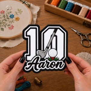 Peut inclure: Un écusson brodé personnalisé représentant le chiffre 10 en blanc avec une bordure noire épaisse. Le motif inclut un club de golf et une balle de golf blanche sur un tee rouge, avec le prénom Aaron brodé en lettres cursives noires sous le chiffre.