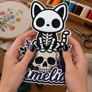 May include: An embroidered patch featuring a black and white skeleton cat sitting atop a beige human skull, surrounded by purple roses and leaves. The word 'Amelia' is stitched in purple script at the bottom of the patch with a white border.