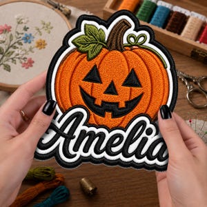 May include: An embroidered patch featuring an orange jack-o'-lantern with a brown stem and green leaf, outlined in black and white. The name Amelia is stitched in a bold, black cursive font with a white border across the bottom of the pumpkin.